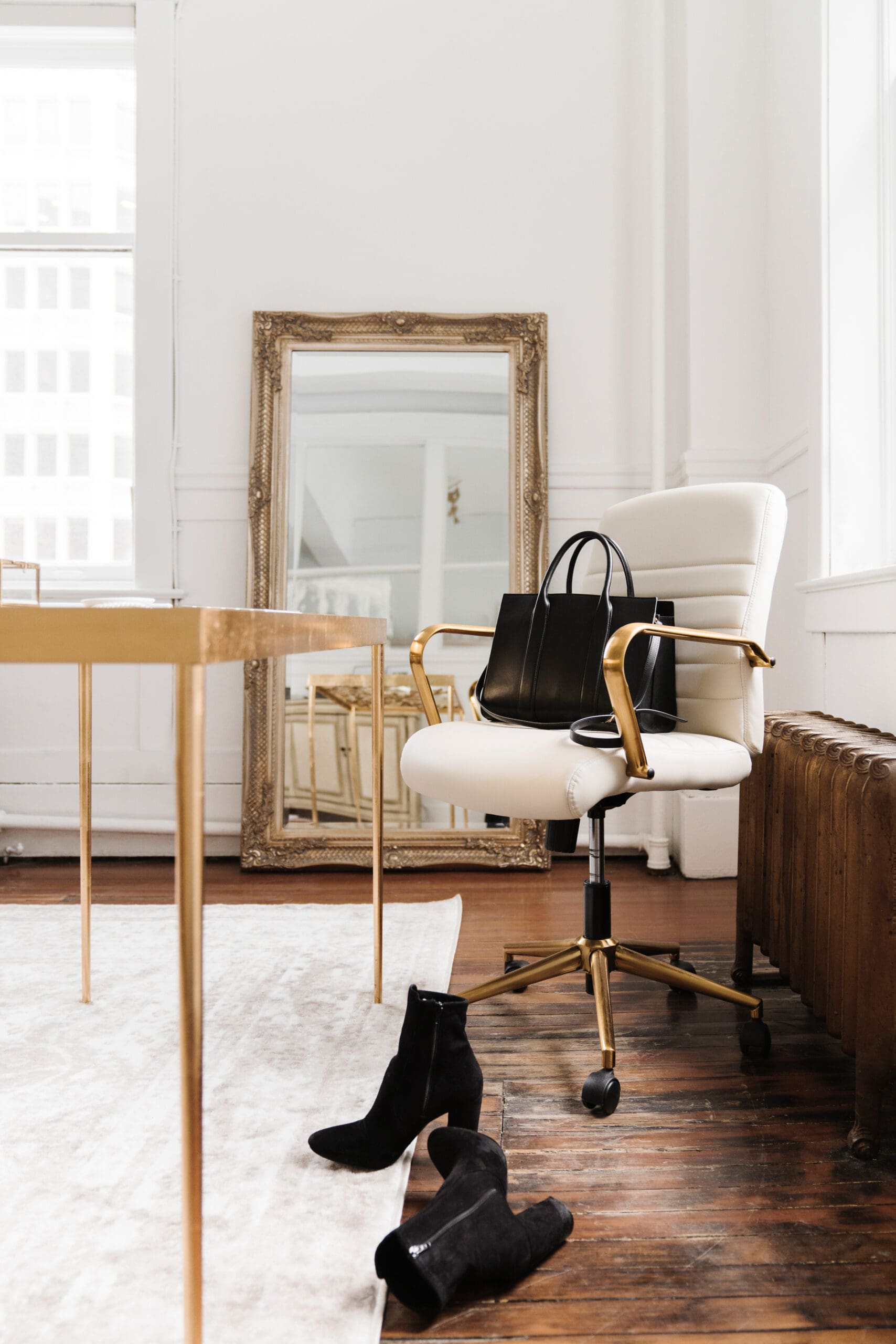 A modern office chair with gold accents and a black tote bag draped over the back, sitting beside a gold desk and large ornate mirror. A pair of black suede boots rest on a white rug, hinting at a moment of pause or transition.