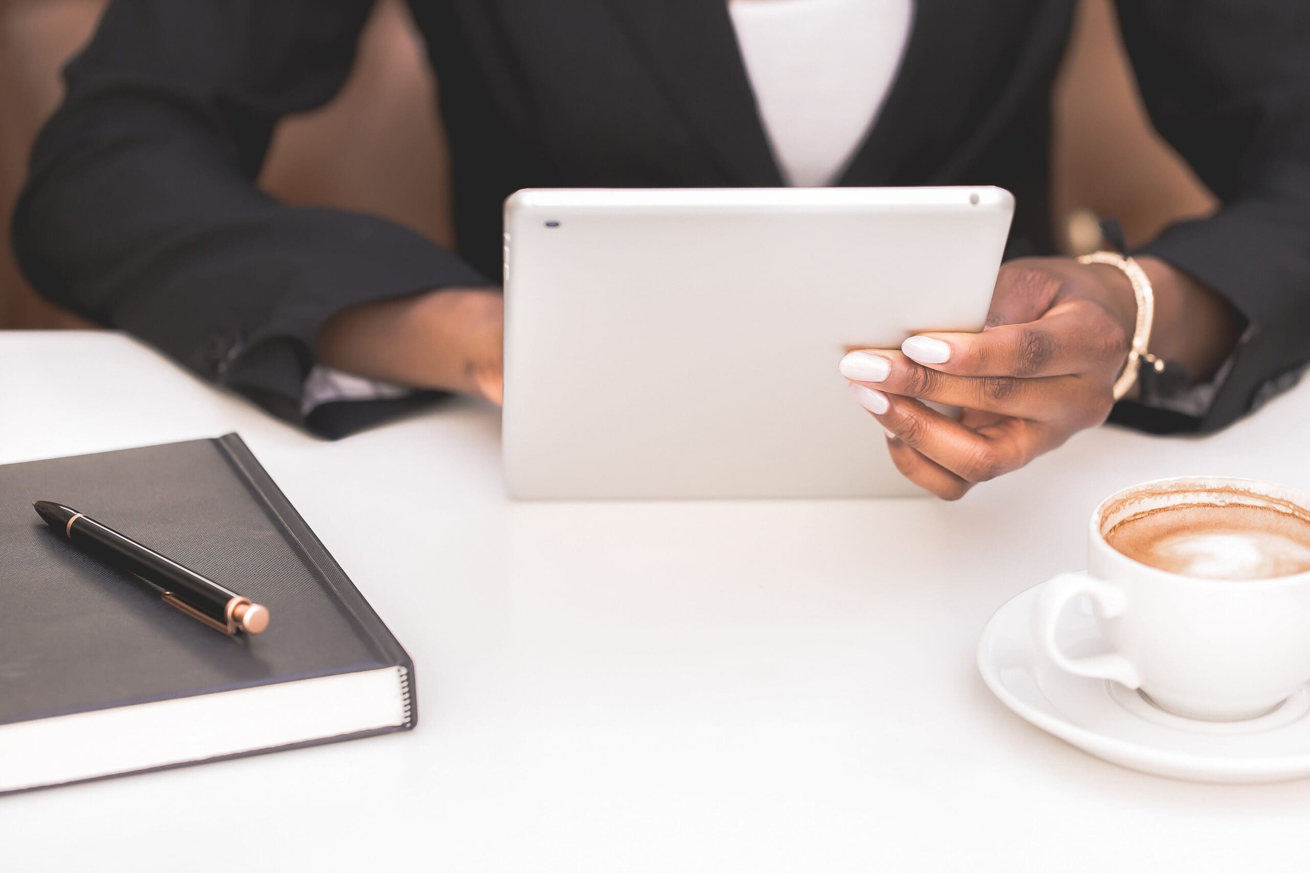 A woman in a black blazer holds a tablet at a white desk with a journal, pen, and cup of coffee nearby—capturing a quiet moment of focused planning or digital work.