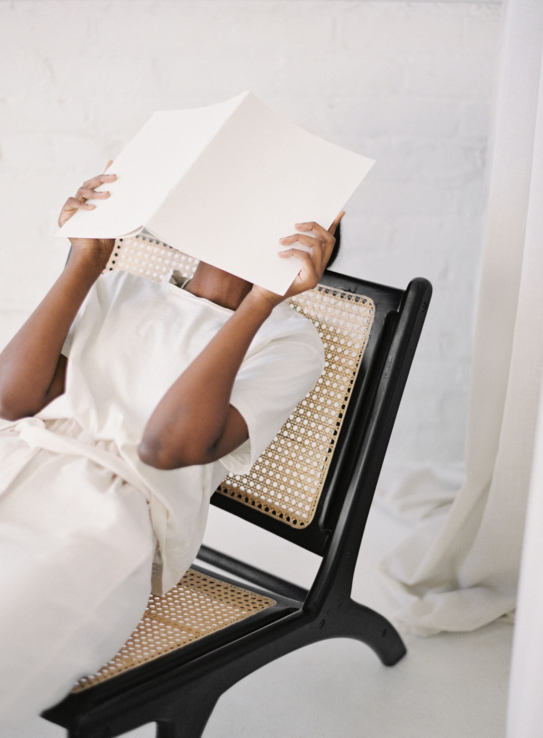 A woman wearing a cream outfit reclines in a woven chair, holding a large white sheet of paper over her face—capturing a moment of creative overwhelm or quiet reflection.