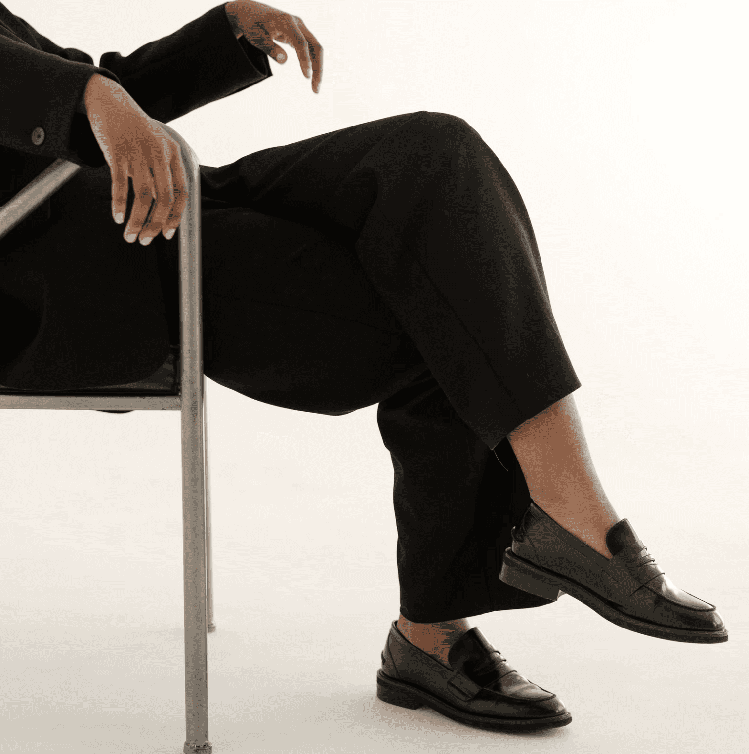 Black woman in tailored black suit sitting confidently on a chair, crossing legs in polished black loafers against a neutral background.