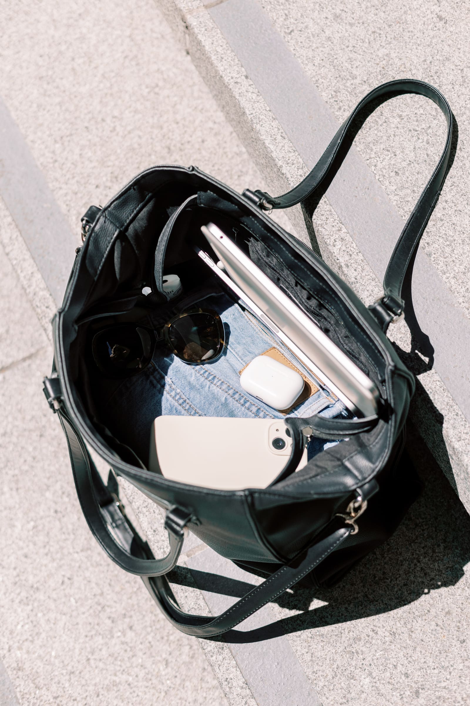 Flat lay of a black leather tote bag on stone steps, filled with both work essentials (laptop, phone) and lifestyle items (sunglasses, denim, AirPods) — symbolizing the balance of soft life and hard skills.